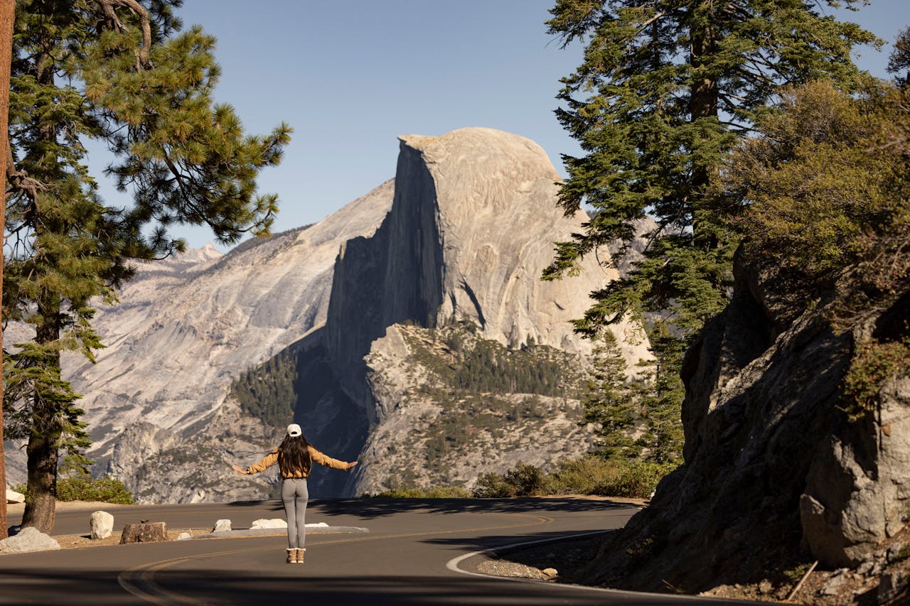 A woman hikes in Yosemite National Park, admiring the iconic Half Dome under a clear fall sky.